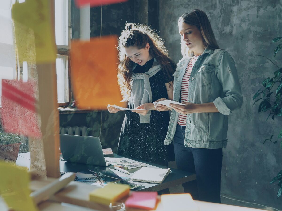 Two colleagues reading printed notes beside a glass wall of sticky notes in a loft office