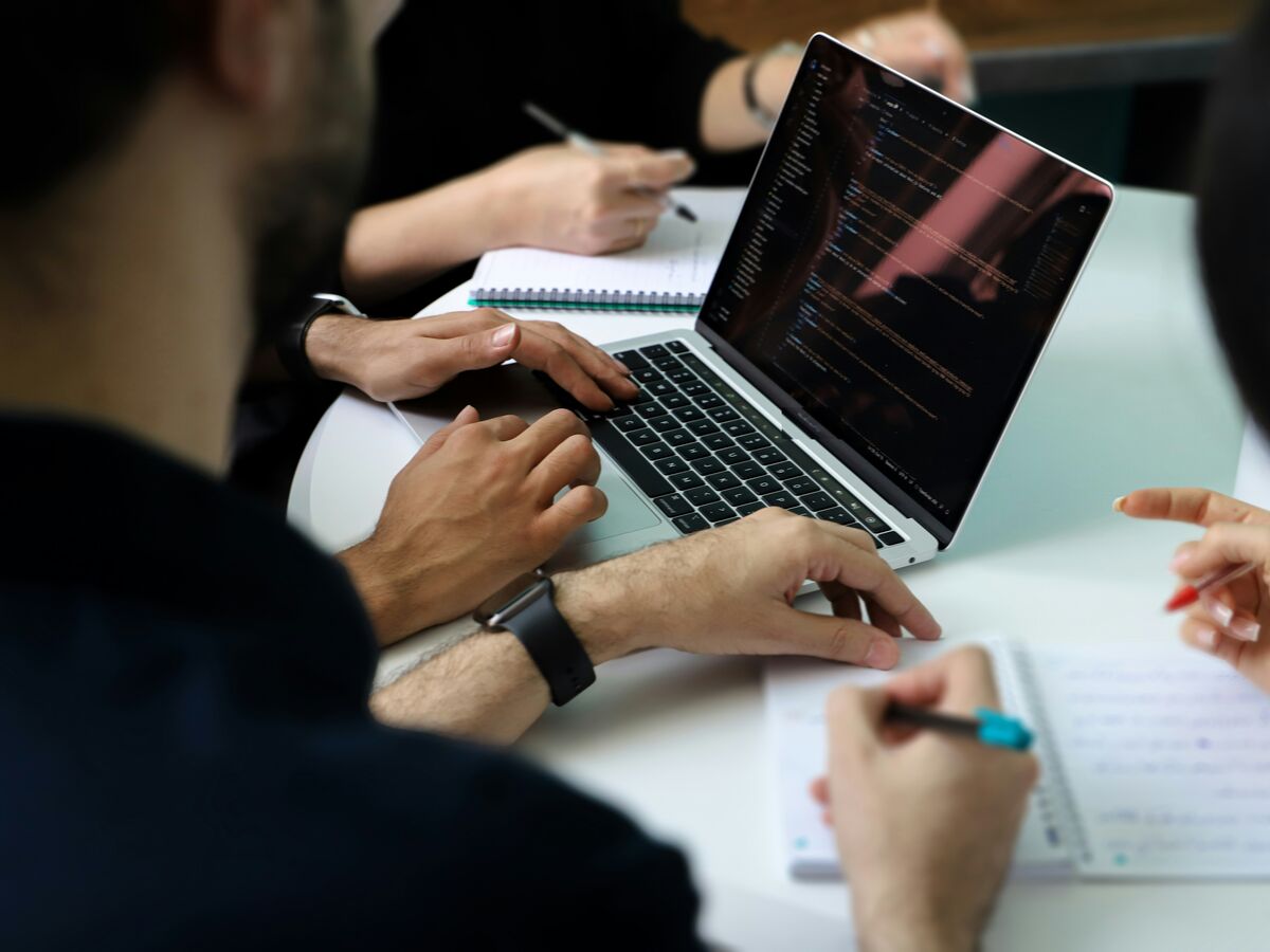 Hands around a laptop showing code, with notes being taken beside it